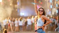 Happy young woman enjoys summer music festival, raises peace sign with sunlit crowd and stage background. Cheerful festivalgoer