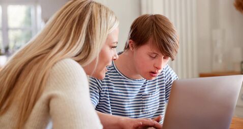 Downs Syndrome Man Sitting With Home Tutor Using Laptop For Lesson At Home
