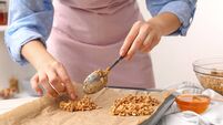 iStock-1937651604.jpg Making granola bars. Woman putting mixture of oat flakes, dry fruits and other ingredients onto baking tray at table in kitchen,