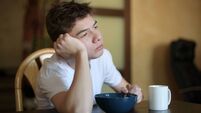 A teenager with a sleepy face idly looks out the window and chews rice porridge