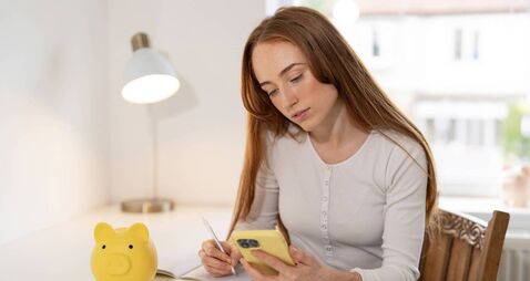Young woman managing finances at home with phone and piggy bank