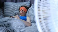 Woman is using eye mask in bed while electric fan is blowing on the foreground