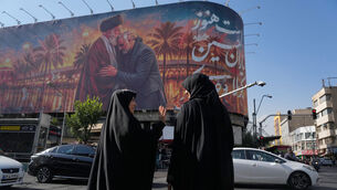 Women talk in front of a banner with graphic depicting the slain Iranian supreme leader Ayatollah Ali Khamenei kissing the head of the late commander of Iran’s Revolutionary Guard expeditionary Quds Force, General Qassem Soleimani, in Tehran, Iran (Vahid Salemi/AP)