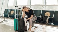 Bored blond woman with luggage, leaning elbow on bags, sit in waiting room at airport due to Covid travel restriction