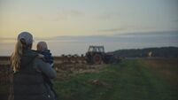 Motherly Farmer Cradles Her Baby Boy,Watching A Tractor Plow The Fields Under The Evening Sky