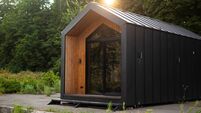 Modern tiny house with wooden and black metal facade, surrounded by greenery, lit by soft evening sunlight.