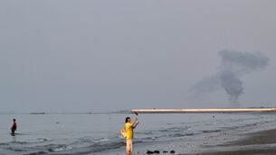 A woman stands at the water’s edge along the Strait of Hormuz as a plume of smoke rises in the background following an explosion, off Bandar Abbas, Iran (Razieh Poudat/ISNA via AP)