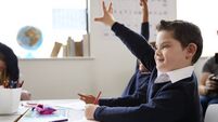 Schoolboy with Down syndrome sitting at a desk raising his hand in a primary school class, close up, side view