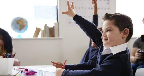 Schoolboy with Down syndrome sitting at a desk raising his hand in a primary school class, close up, side view