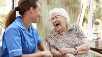 Senior Woman Sitting In Chair And Laughing With Nurse In Retirement Home