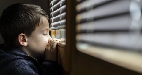 Small sad boy looking through the window during Coronavirus isolation.