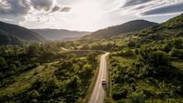 iStock-2279600441.jpg Car driving on winding road through lush green valley at sunset