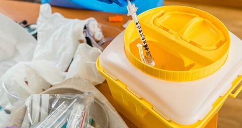 iStock-1299861506.jpg Hand with blue latex glove throws syringe into yellow waste container in hospital