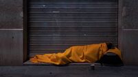 A homeless person sleeps on the street under a blanket in front of a storefront