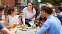 Waitress Serving Drinks To Group Of Friends Sitting At Table In Pub Garden Enjoying Drink Together