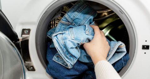 Person putting jeans into the drum of a washing machine, front view. Washing dirty jeans in the washer