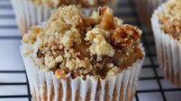 Close-up image of rows of homemade apple muffins in paper cake cases topped with crumble on wire metal cooling rack, marble effe