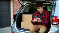 Young man texts on phone while sitting in car trunk with boxes. He is moving out of home for college, starting new life journey.