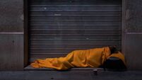 A homeless person sleeps on the street under a blanket in front of a storefront