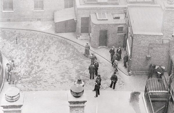 In the prison courtyard of the Bow London Court during the recess hour of the Casement Trial. On the extreme right is Roger Casement, seated with a sheet of paper in his hand. On the left, with his back to the camera, Bailey, alleged co-conspirator with Casement. In the background with his left arm behind his back is Corporal O’Connor. On the top right is Mary Gorman, the Kerry Collen who aided in capturing Casement. Mid-stride directly in front of the London bobby on the right is Martin Collins.
