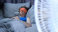Woman is using eye mask in bed while electric fan is blowing on the foreground