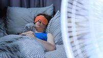 Woman is using eye mask in bed while electric fan is blowing on the foreground