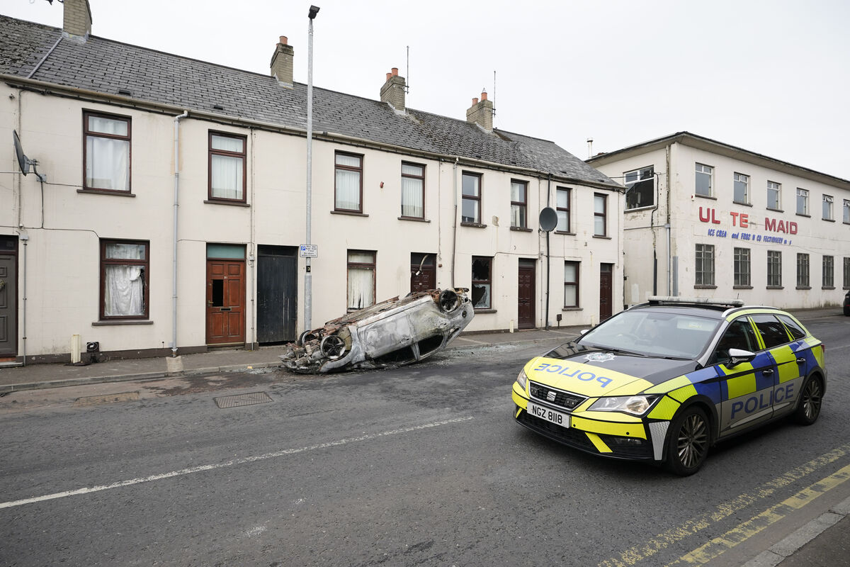 A police car drives past a burnt-out overturned car on the street following a second night of violence in Ballymena.