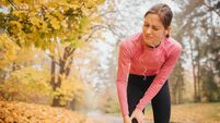 Slim and well-built young woman stands on road in autumn park. She holds hands on knee. Model feels pain there. She suffer.
