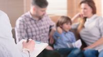 Hand of a professional family psychotherapist writing notes in front of a couple with a child in a blurred background during a c