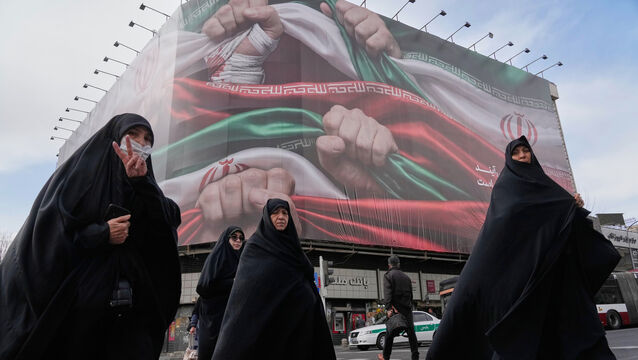 Women cross a street under a huge banner showing hands firmly holding Iranian national flags – a sign of patriotism – in Tehran, Iran (Vahid Salemi/AP)