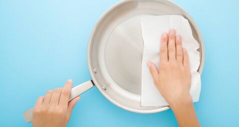 Young adult woman hand wiping stainless frying pan with dry white paper napkin on light blue table background. Closeup. Pastel c
