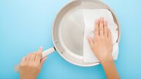 Young adult woman hand wiping stainless frying pan with dry white paper napkin on light blue table background. Closeup. Pastel c
